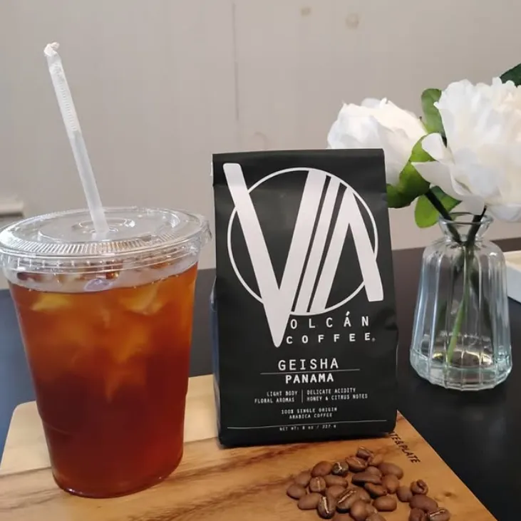 Black Vía Volcán Geisha Panama coffee bag next to an iced coffee and white flowers