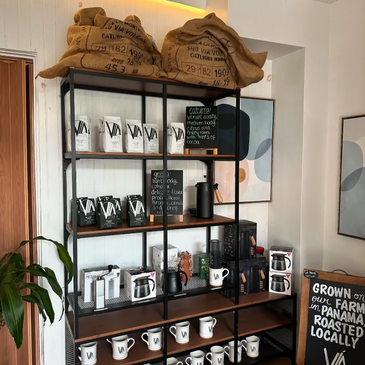 Shop shelf stocked with Vía Volcán coffee bags, branded mugs, and burlap coffee sacks, beside a chalkboard reading "Grown on our farm in Panama, roasted locally"